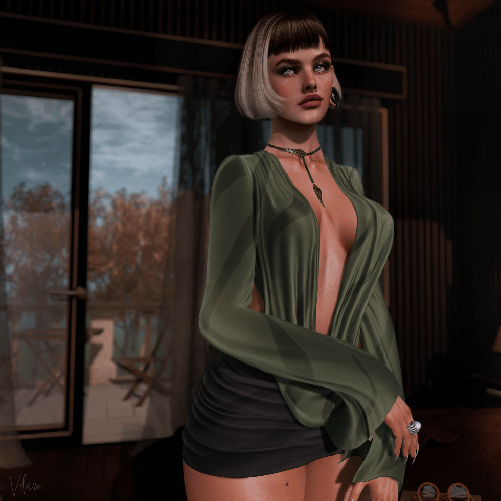 A stylish woman with a sleek blonde bob and dark bangs poses in a dimly lit, elegant room, wearing an olive-green blouse, a black skirt, and delicate jewelry. Her striking gaze and composed expression create a mysterious, sultry mood, while warm lighting and autumn foliage outside the windows add to the cinematic atmosphere.